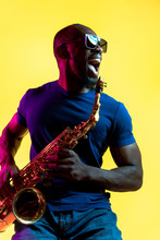 Young African-american Jazz Musician Playing The Saxophone On Yellow Studio Background In Trendy Neon Light. Concept Of Music, Hobby. Joyful Attractive Guy Improvising. Colorful Portrait Of Artist.