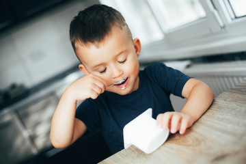 Wall Mural - Cute boy in the kitchen eating yogurt from a white container for yogurt, there is a place for advertising