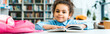 © LIGHTFIELD STUDIOS - panoramic shot of happy african american kid sitting near book in library
