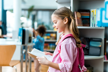 cheerful kid with pink backpack standing and reading book in library