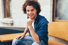 Smiling Smart Handsome Young Man With Curly Hair, Looking To The Camera And Sitting On The Bench In The City Street. Happy Male Student Relaxing Outdoors. People And Lifestyle Concept