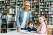© LIGHTFIELD STUDIOS - cheerful teacher in glasses standing near cute kids in library