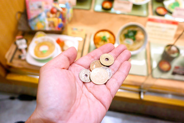 Image from eyes looking Japan Yen coins on hand front of blurry Japanese restaurant display, It shows that the cost of living in Japan is so high and the price of food in restaurants is high too.