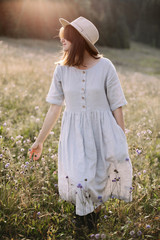 Stylish girl in rustic dress walking in wildflowers in sunny meadow in mountains. Boho woman relaxing in countryside flowers at sunset, rural simple life. Atmospheric image. Space text