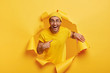 Positive young male model stands in hole of ripped paper background, points at himself, asks about choosing his own canditature, wears yellow hat and t shirt, being in high spirit, smiles broadly