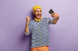© WHstudio Leushin N - Discontent emotional angry man takes selfie picture, expresses negative emotions on camera, raises clenched fist, wears yellow hat and striped jumper, isolated on purple background, uses headphones