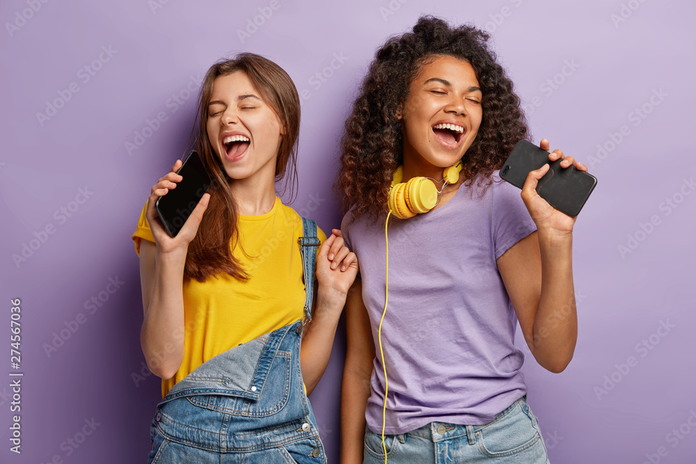 Optimistic pleased mixed race women sing favourite song in smart phones, have fun and enjoy music, keep eyes shut, move actively, isolated on purple background. People, free time and joy concept - obrazy, fototapety, plakaty 