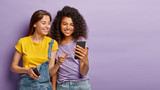Fototapeta  - Pleased female friends watch something interesting on mobile phone, have happy relaxed expressions, focused in device, enjoy using modern technologies, isolated over purple wall with empty space