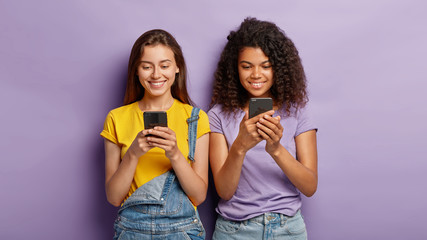 Two mixed race beautiful women send text messages on mobile phones, being addicted to modern technologies, make shopping online, have glad facial expressions, stand next to each other on purple wall