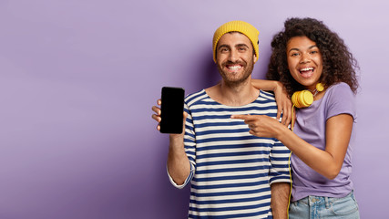 Selective focus of happy woman and man points to modern device with blank screen for your advertising, promot cool gadget, have smiles, wear stylish outfit, stands over purple wall, free space on left