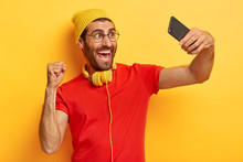 Triumphing Glad Man Celebrates Success, Makes Photo Of Himself, Takes Selfie, Shoots Video, Wears Hat, T Shirt And Spectacles Isolated Over Yellow Background. People, Lifestyle, Spare Time, Technology