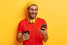 Half Length Shot Of Positive Guy Smiles At Screen Of Smartphone, Has Online Conversation In Chat, Forgets About All Problems, Holds Takeaway Coffee, Dressed In Red T Shirt, Yellow Hat On Head