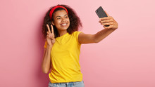 Lovely Young Curly Female Poses In Smartphone Camera, Makes Peace Gesture, Takes Photo For Social Networks, Enjoys Free Time, Dressed Casually, Being On Way To Shop, Isolated On Pink Background