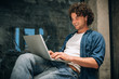 © iuricazac - Portrait of happy young man with curly hair using laptop for working online. Smart freelance man chatting online with clients. Handsome male reading news, typing on his laptop computer in the office