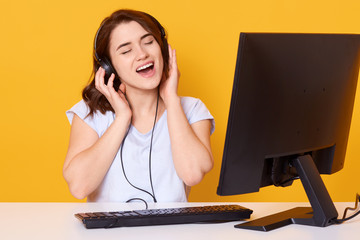 Studio shot of girl listening music with headphones and singing songs loudly, sitting at white desk in front of personal computer, lady dressed casual white t shirt, posing against yellow studio wall.