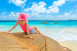 Seychelles, Anse Lazio, Indian Ocean. Young tourist woman with pink hat, relaxing on a large boulder typical of the most famous beach of Praslin.Female lifestyle looks at the horizon and turquoise sea