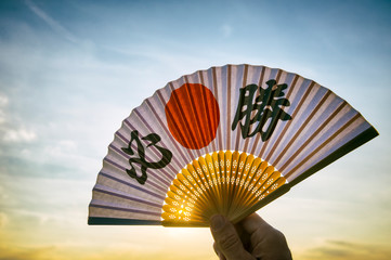 Hand of Japanese sports supporter holding a fan decorated with kanji characters spelling out hissho (English translation: certain victory) at sunrise