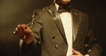 Male Orchestra Conductor Wearing Tux Standing In Front Of Music Stand, Controlling Musicians By Moving His Hands And Baton. Studio Shot On Black Background 4k Footage