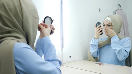 Wall Mural - Muslim young attractive woman in beige hijab and traditional blue dress making make-up and standing in front of the mirror.