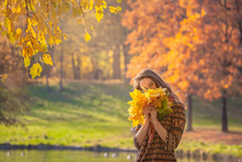A Young Woman Walks In The Autumn Park, She Stands By The Lake, Brunette Woman Wearing A Grey Coat, She Is Holding A Bouquet Of Yellow Leaves