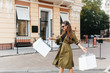 © Look! - Pleased woman with long dark-brown hair walking down the street and waving bags from boutique with smile. Outdoor photo of inspired shopaholic lady in sunglasses enjoying weekend in autumn.