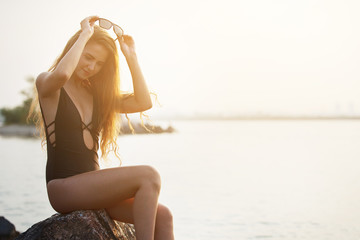 Wall Mural - Young, pretty woman in a black swimsuit is sitting on the rocks by the sea on a sunset background.