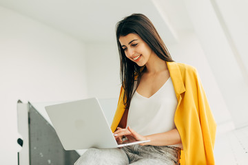 Wall Mural - Busy cheerful young woman work in white room. Hold laptop on knees and typing on keyboard. Smiling look at screen. Remote work on project. Sit in coworking space.