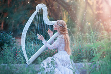 Mysterious Forest Nymph Plays On White Harp In Fabulous Place, Girl With Long Blond Hair And Elegant Lace Vintage Dress Calling For Bright Sun Rays, Lady With Silver Jewelry And Musical Instrument.