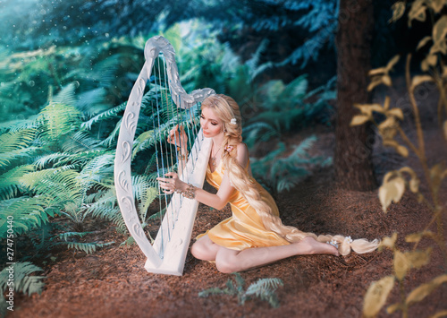 Fotografía fabulous forest elf sits under tree and plays on white harp, girl with long blond hair braided in long yellow dress, summer goddess rests and sings to the sound of a magical musical instrument