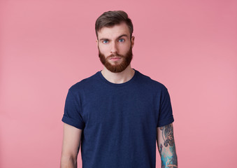 Wall Mural - Photo of young handsome tattooed misunderstanding red bearded man in blank t-shirt, stands over pink background, looks at the camera with raised eyebrow.