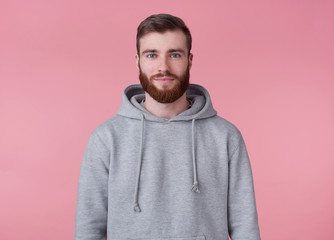 Wall Mural - Portrait of young attractive red bearded man in gray hoodie, looks good and calm, smiles, stands over pink background.