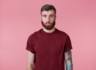 Wall Mural - Portrait of sad young handsome red bearded tattooed man in red t-shirt, offended looking at the camera with a lowered lip, stands over pink background.