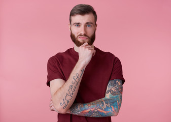 Wall Mural - Portrait of young handsome thinking tattooed red bearded man in red t-shirt, touches the chin, stands over pink background looks at the camera.