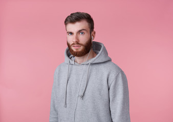 Wall Mural - Portrait of young attractive red bearded man in gray hoodie, looks good and calm, smiles, istening song in headphones, enjoys the music, stands over pink background.