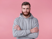 Portrait Of Young Frowning Handsome Red Bearded Man In Gray Hoodie, Stands With Crossed Arms, Disapprovingly Looks At Camera, Stands Over Pink Backround.