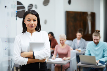 Waist up portrait of female teacher smiling at camera while standing by whiteboard with group of students in background, copy space