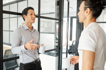 Young Asian businessman with cup of coffee explaining idea to coworker during short break