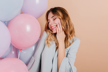 Romantic curly girl in blue attire posing with eyes closed and sincere smile. Indoor photo of carefree white lady celebrating birthday with balloons.