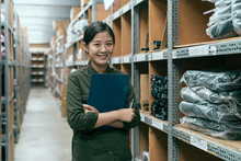 Cute Female Worker Holding Clipboard In Warehouse. Young Attractive Girl Staff Standing In Storehouse Face Camera Smiling. Happy Woman Stockroom Employee Surrounding By Goods Inventory In Factory.