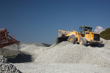 Wheel loader excavator against the background of gravel hills and blue sky at a crushing and sorting factory, close-up. Mining industry. Quarry mining equipment.