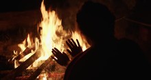 Faceless Woman Warming Up By Campfire Late At Night. Unrecognizable Tourist Girl Holding Hands Close To Fire Outdoor Nature Black Background Back View Slow Motion. Female Camping Hiking Danger Concept