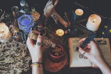 A Witch Casting A Spell And Writing It Down In Her Book Of Shadows With A Green Black Feather And Ink. Wiccan Witch Altar Filled With Nature Objects Branches Dried Flowers Herbs Burning Candles Potion