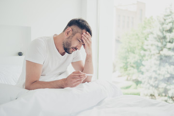 Close up side profile photo amazing he him his unhappy guy hold thermometer terrible hurt pain illness temples forehead need go work job white sleeping wear sit big large bedroom room house indoors