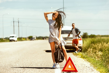 The young couple broke down the car while traveling on the way to rest. They are trying to stop other drivers and ask for help or hitchhike. Relationship, troubles on the road, vacation.