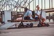 © Fxquadro - Three good happy friends are sitting on the bench at riverside. They have bagpacks and electronic scooters.