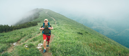 Wall Mural - smiling hiker man walking by the foggy cloudy weather mountain range path with backpack. active spor