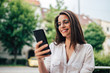 © bnenin - Portrait of a happy elegant millennial woman looking at smart phone outdoors.