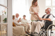 © Photographee.eu - Senior man on wheelchair with helpful nurse holding his hand and friends sitting on couch drinking tea