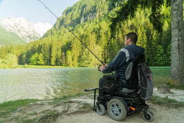 Wall Mural - young man in a wheelchair fishing at the beautiful lake on a sunny day, with mountains in the back