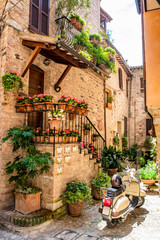 A characteristic alley of the medieval village, with stone and brick houses, plants and flowers on the balconies. A wasp parked in the street. In Spello, province of Perugia, Umbria, Italy.